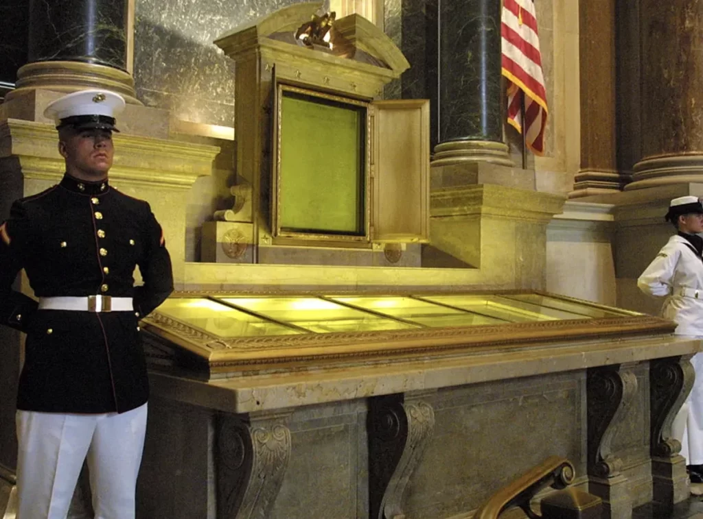 Honor guard standing beside the original display cases of the Declaration of Independence and the U.S. Constitution at the National Archives, with the American flag visible in the background.