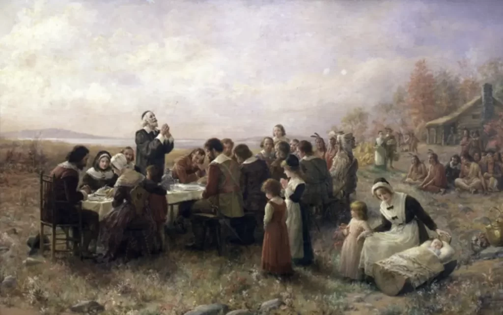 A painting by Jennie Augusta Brownscombe titled "The First Thanksgiving" from 1914, housed at the Pilgrim Hall Museum in Plymouth, MA, USA. The artwork depicts Pilgrims and Native Americans gathered outdoors around a large table for a shared meal, surrounded by autumn foliage and a rustic cabin. A woman is seated with children nearby, adding a serene, communal atmosphere. ​