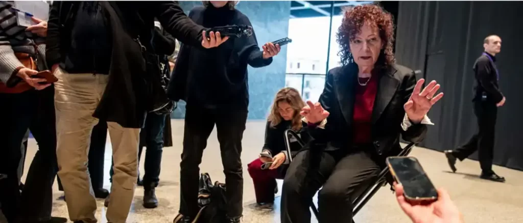 Nan Goldin seated and gesturing during a press interaction at the Neue Nationalgalerie, surrounded by reporters holding recording devices and microphones.