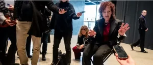 Nan Goldin seated and gesturing during a press interaction at the Neue Nationalgalerie, surrounded by reporters holding recording devices and microphones.