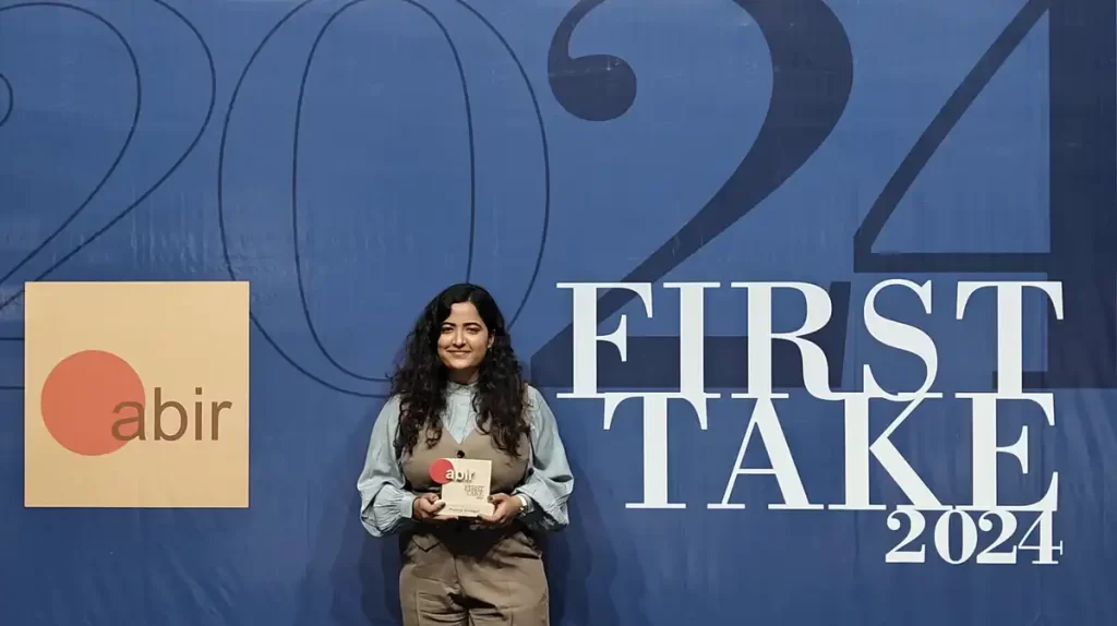 Pushpa Kumari holding her First Take 2024 award, standing in front of a backdrop featuring the First Take 2024 logo and the abir brand.