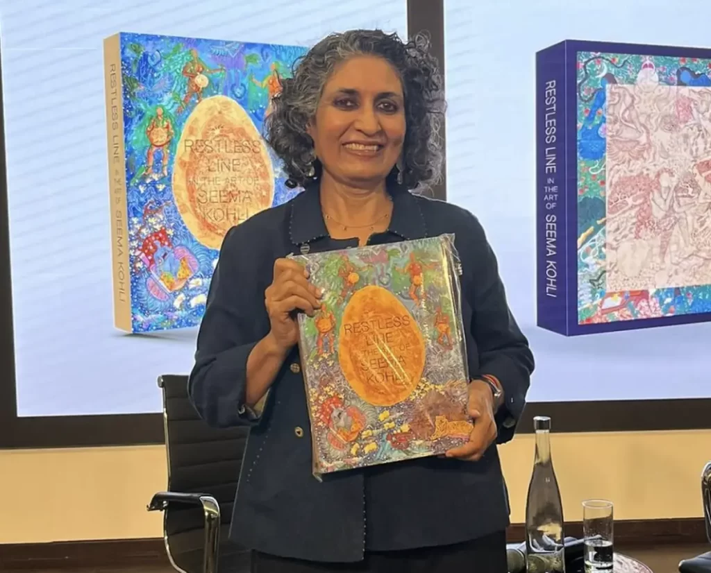 Seema Kohli proudly holding her book Restless Line in the Art of Seema Kohli during its release event, with vibrant artwork from the book displayed in the background.