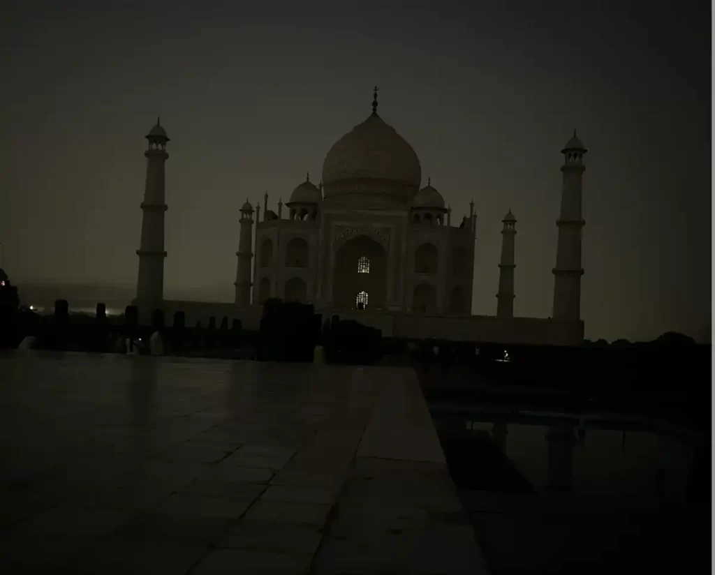 A serene view of the Taj Mahal captured at dawn, with the iconic white marble structure and its minarets silhouetted against a dimly lit sky. The soft lighting enhances the monument's timeless beauty, reflecting a tranquil and peaceful atmosphere