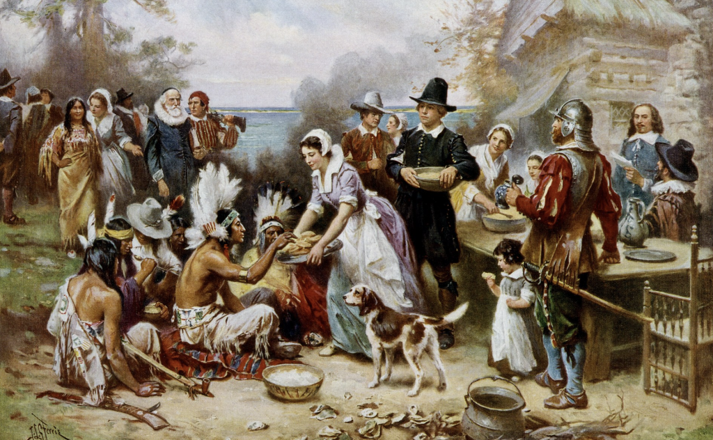 An artistic depiction of the first Thanksgiving, showing a harmonious gathering of Native Americans and Pilgrims sharing food outdoors. The image features Native Americans seated on the ground wearing traditional feathered headdresses, while Pilgrims in colonial attire, including women in bonnets and men in armor or dark clothing, serve food from bowls and plates. A dog is present in the foreground, and a wooden cabin with smoke rising from a chimney is visible in the background, suggesting a communal and celebratory atmosphere.