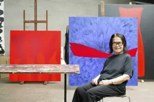 Tomie Ohtake seated in her studio, wearing black clothing and glasses, smiling gently. Behind her are vibrant abstract paintings, including a red canvas with subtle tonal variations and a blue canvas featuring a bold red curved form, reflecting her signature style of abstract expressionism.