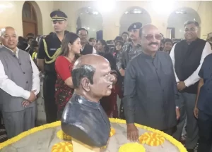 A ceremonial gathering featuring a bust sculpture of a notable figure surrounded by marigold garlands. People, including officials and guests, stand around, engaging in conversation in a formal indoor setting.