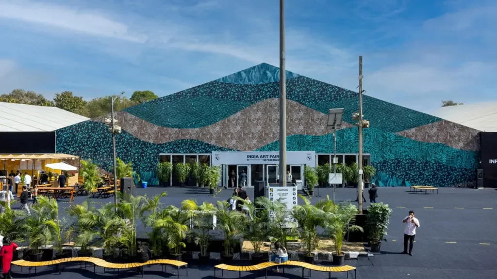 Exterior view of the India Art Fair entrance featuring a vibrant and intricate mural with blue, green, and brown patterns, surrounded by lush potted plants and benches. Visitors are seen entering and exploring the area under a bright, clear sky.