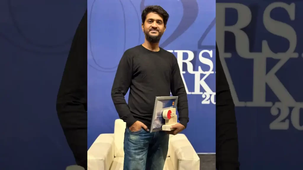 Sandipan Acharjee, one of the winners of Abir First Take 2024, holding his award while standing in front of the event backdrop, wearing a black shirt and blue jeans, smiling confidently.